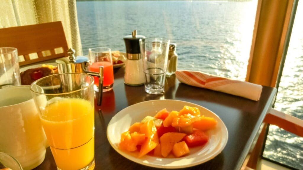A breakfast on a cruise with vibrant fruit and orange juice is set against a stunning sea view, basking in the warm morning sunlight.