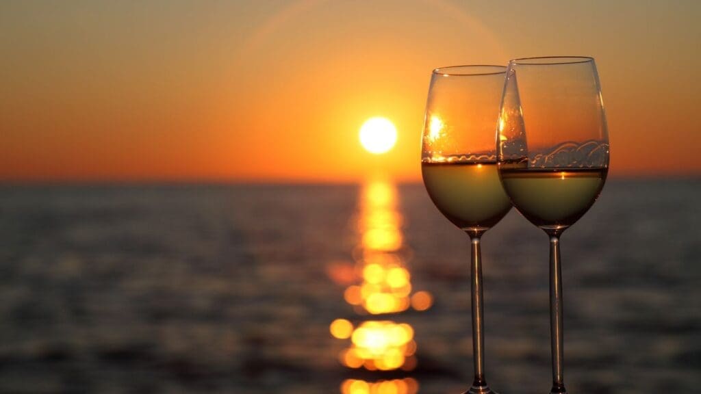 Two glasses of white wine glisten in the warm glow of a vibrant sunset over the ocean, creating a serene and romantic cruise atmosphere.