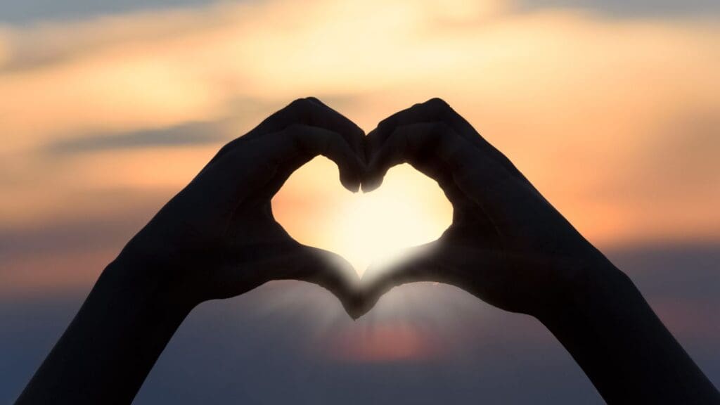 Hands form a heart shape around the setting sun, creating a silhouette against a vibrant evening sky on a cruise.
