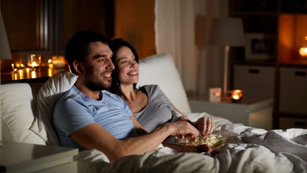 A couple enjoys a romantic private movie night in bed, surrounded by the warm glow of candlelight, sharing a bowl of popcorn on a cruise.