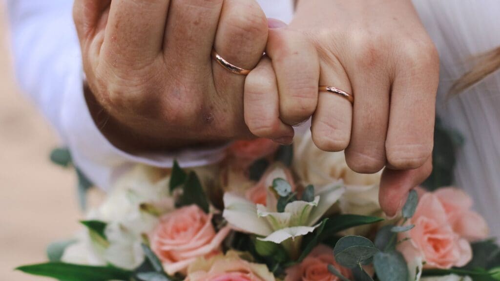 Newlyweds celebrate a vow renewal on a cruise with a touching pinky promise, their wedding rings gleaming against a backdrop of delicate roses and greenery.