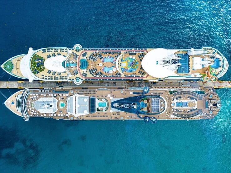 Aerial view of two luxurious cruise ships docked side by side, showcasing vibrant decks filled with pools, sports courts, and leisure facilities against the deep blue ocean.